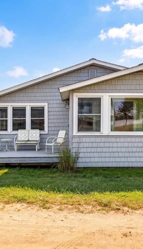 Gray shingled cottage with small front deck and white patio chairs on a sunny summer day